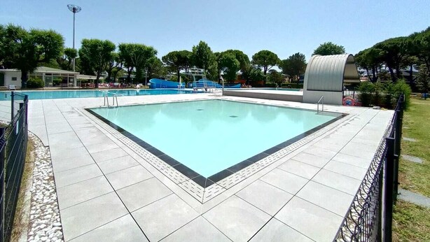 A rectangular swimming pool with a tiled deck, surrounded by a metal fence. - Appartamenti Olimpia (San Michele al Tagliamento)