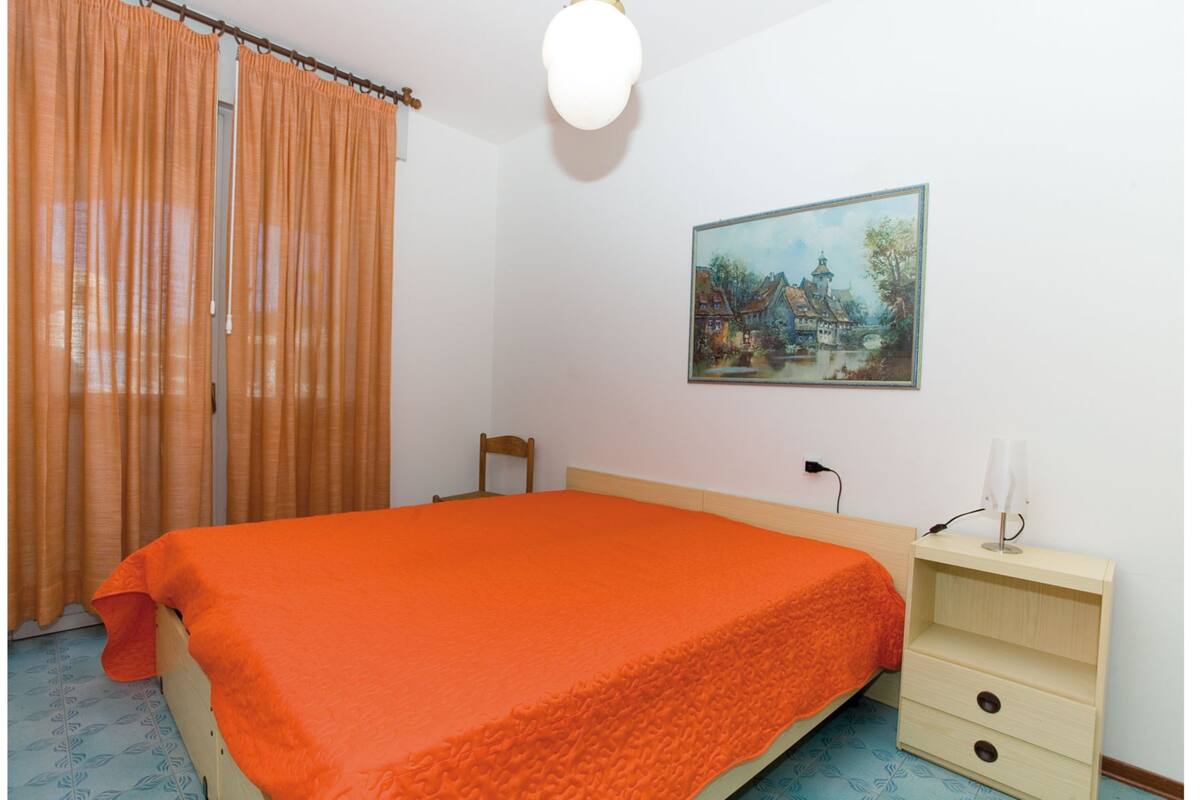 Une chambre à coucher avec un lit, une table de chevet, une chaise, un tableau au mur et une fenêtre avec des rideaux.