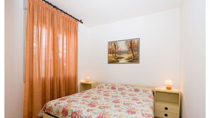 A bedroom with a floral bedspread, a painting of a forest path on the wall, and two bedside tables with lamps. - Appartamenti Olimpia (San Michele al Tagliamento)