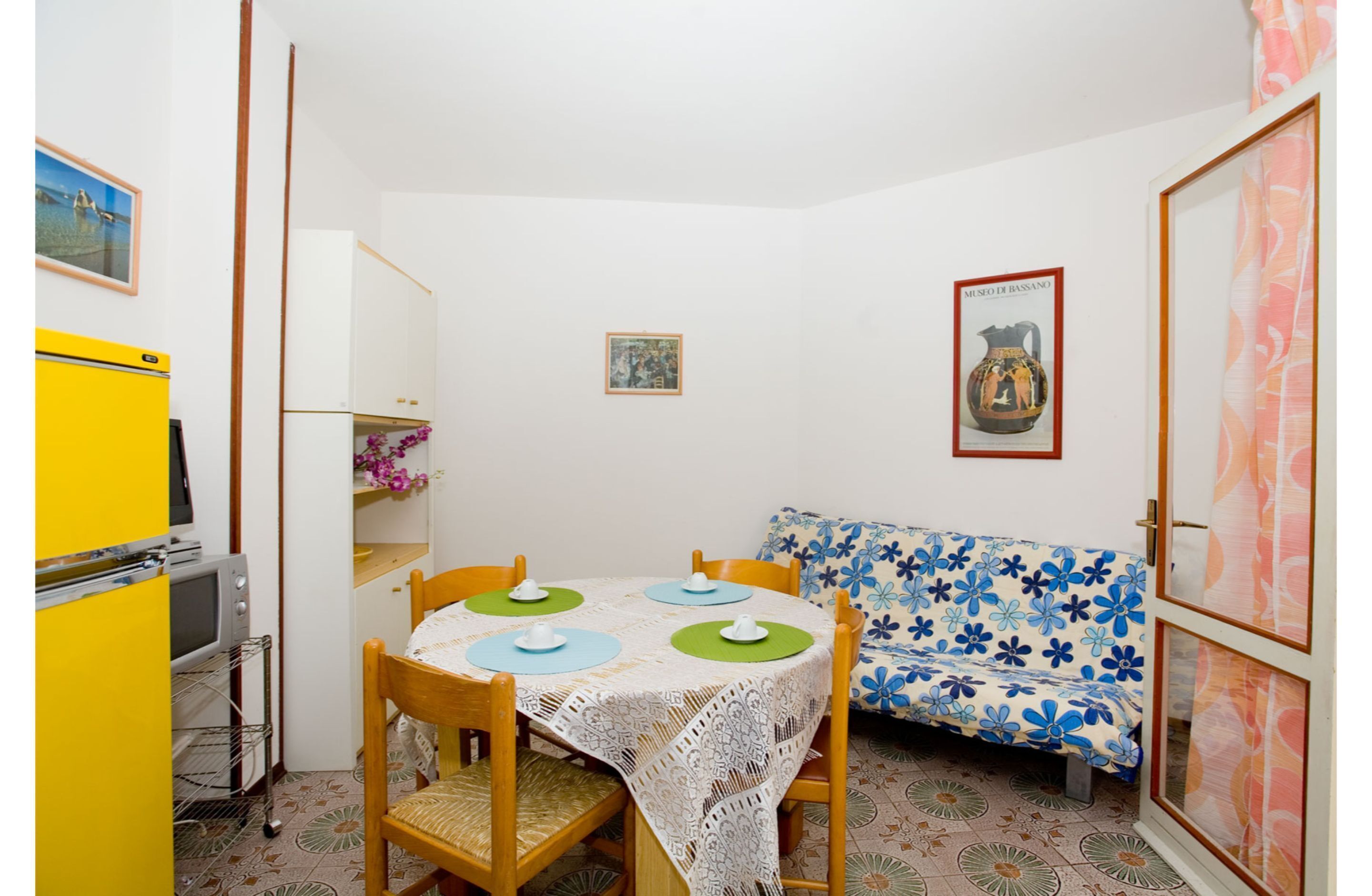 A compact room with a dining table, chairs, a sofa, a small refrigerator, and a television.