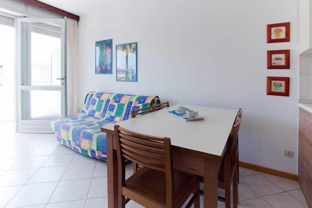 A dining area with a white table, wooden chairs, a colorful sofa, and a sliding glass door.
