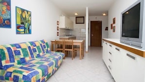 A compact living space with a colorful sofa, a dining area with wooden chairs, a kitchen with white cabinets, and a large TV on the right.