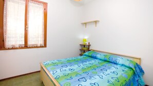 Een kleine slaapkamer met een bed, een raam met kantgordijnen, een nachtkastje met een lamp en een houten plank.