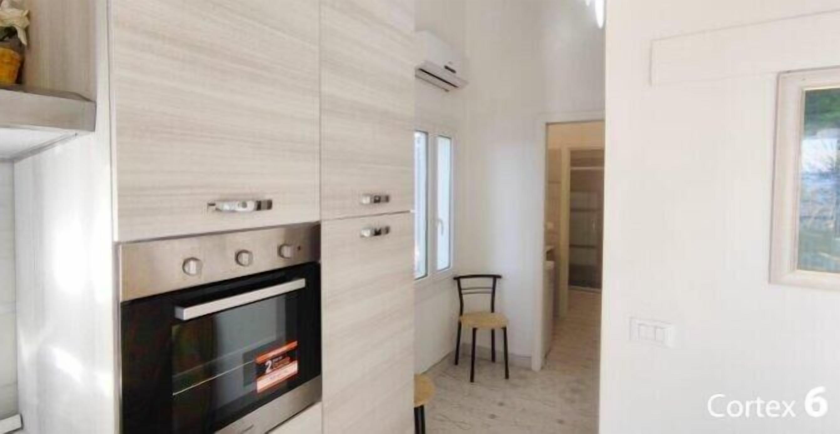 A modern kitchen with built-in appliances, a small dining area with a chair, and a view of the outside through a window.