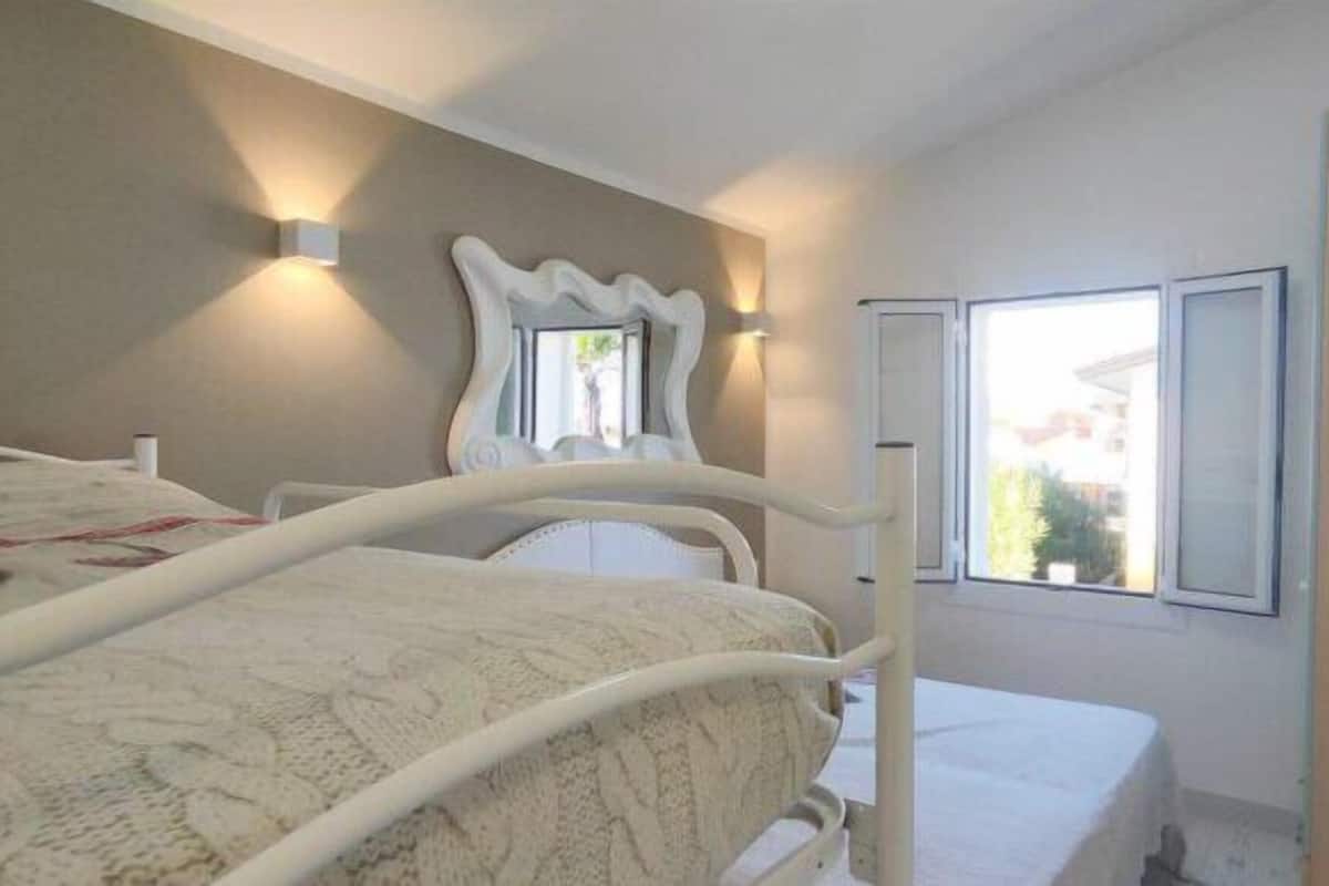Un dormitorio con cama, mesita de noche, espejo y ventana con vistas a edificios.