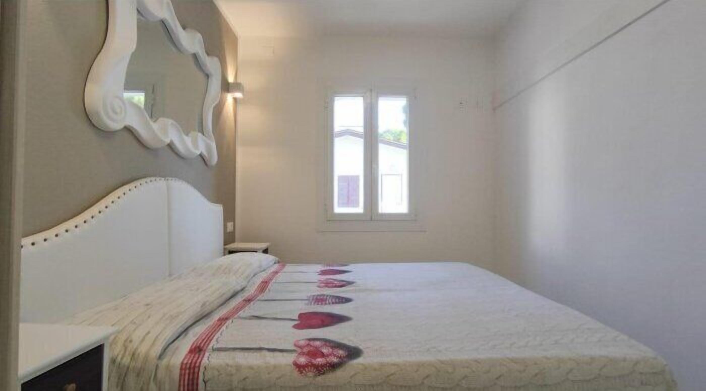 Un dormitorio con cama, espejo, ventana y mesita de noche.