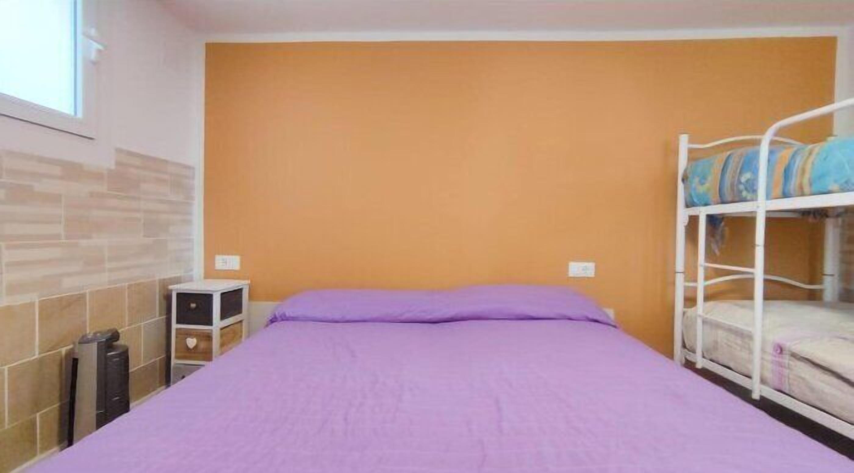 Una cama individual con una colcha morada en una habitación con pared naranja y una cama litera a la derecha.