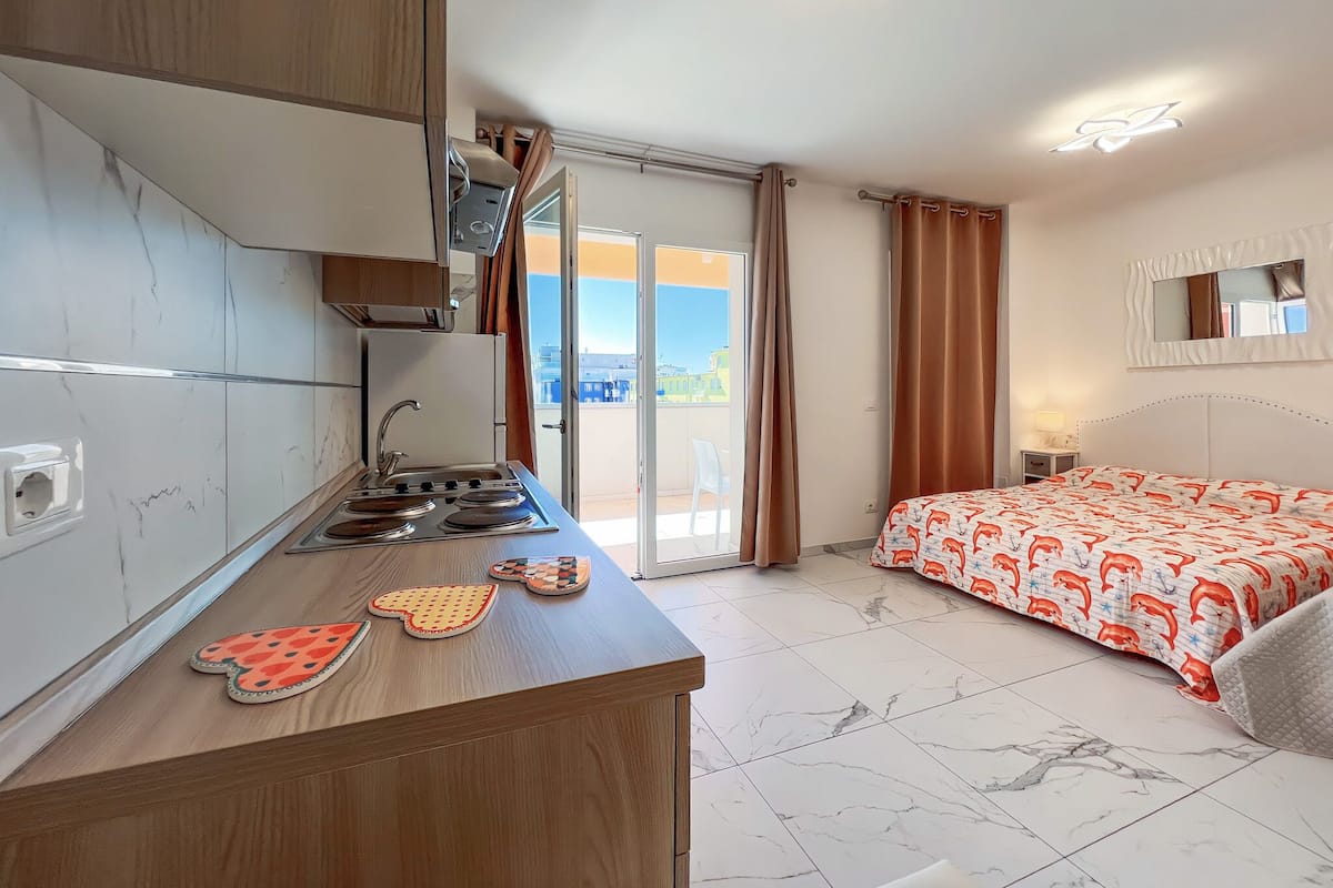 Een compacte hotelkamer met kitchenette, een bed met een bedovertrek met dessin, een groot raam met gordijnen en een plafondlamp.