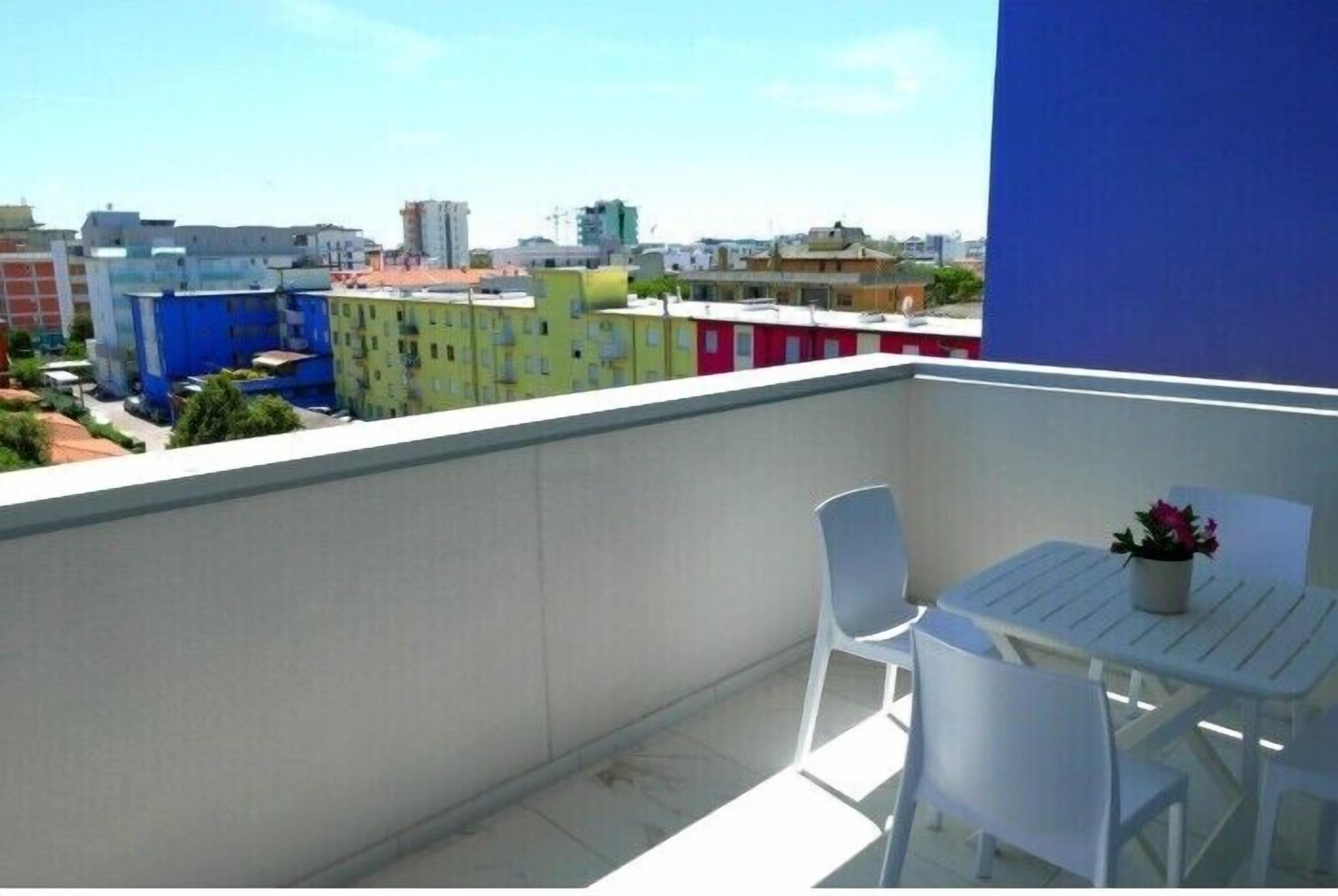 A balcony with white chairs and a table, overlooking a cityscape with colorful buildings.