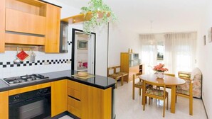 A compact kitchen with wooden cabinets, a black countertop, and a built-in oven. There is a dining area with a wooden table and chairs, and a hanging plant above the kitchen counter. - Appartamenti Tiziano (San Michele al Tagliamento)