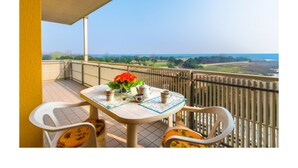 Un balcon avec une table dressée pour deux personnes, des chaises et une vue sur la mer et la verdure.