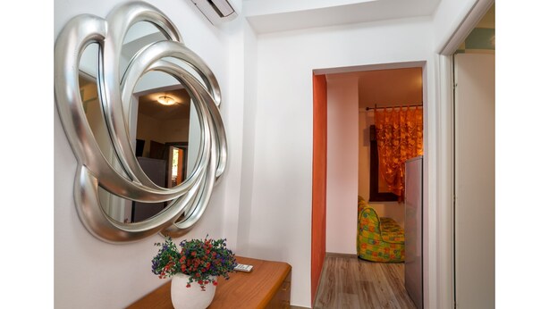 A hallway with a wall-mounted mirror, a wooden console table with a potted plant, and a colorful sofa in the background. - Appartamenti Krystall (San Michele al Tagliamento)