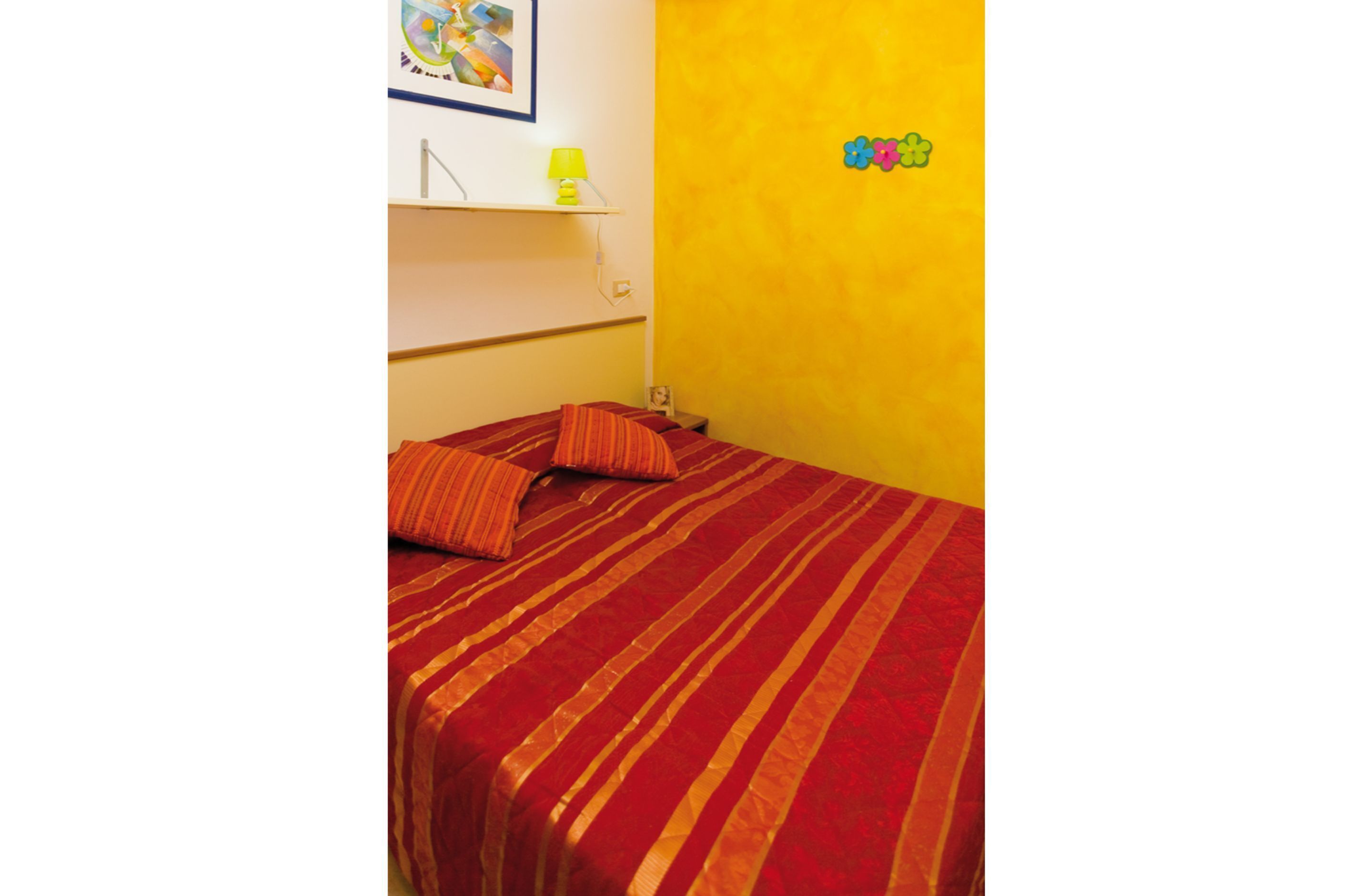 A room with a bed covered in a striped red and orange bedspread, a small shelf with a lamp, and a wall with a flower decoration.