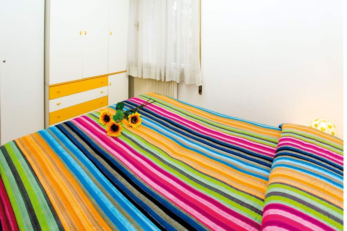 A room with a bed featuring a colorful striped bedspread, a wardrobe with yellow accents, and a window with white curtains.