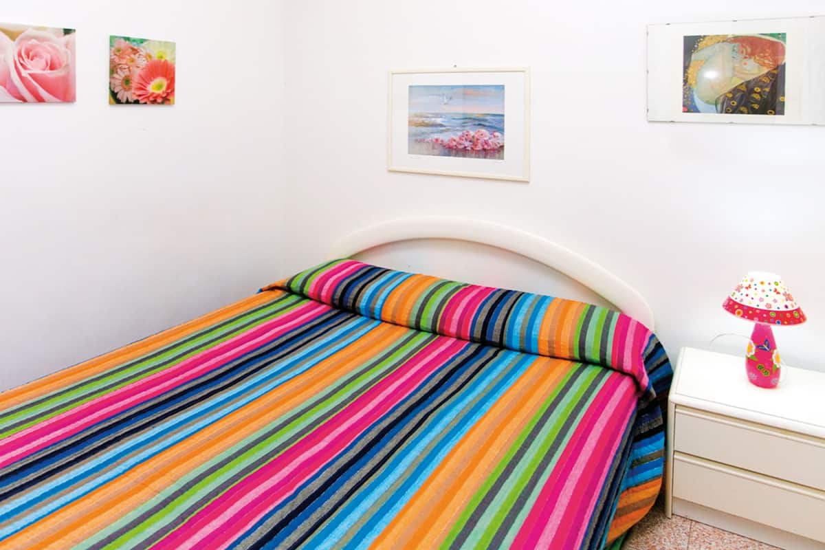 A bedroom with a colorful striped bedspread, a white curved headboard, a small bedside table with a lamp, and three framed pictures on the wall.