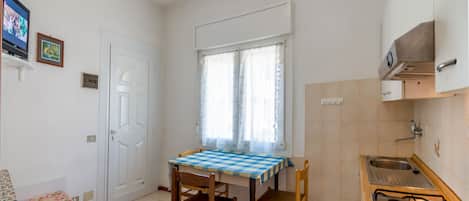 A compact kitchen with a gas stove, a sink, and a window with curtains. There is a table with chairs and a television on the wall.