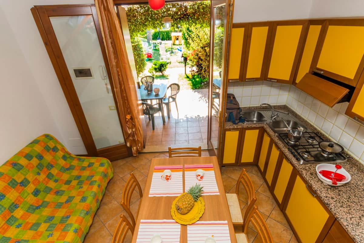 Une cuisine compacte avec un coin repas, une petite table ornée d’un ananas, et une vue sur le jardin par la porte ouverte.