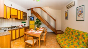 Eine kleine Küche mit gelben Schränken, ein Esstisch mit Stühlen, eine Holztreppe und ein farbenfrohes Sofa.