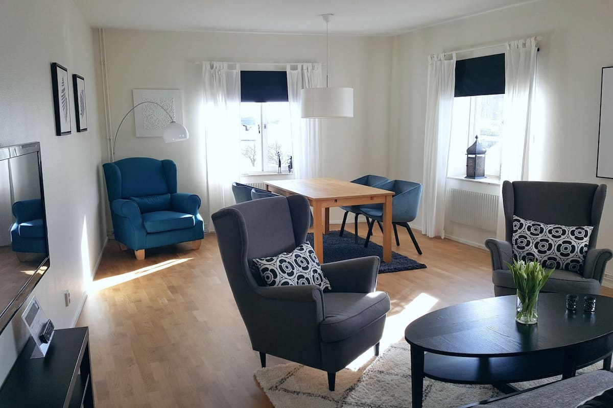 A modern living room with a wooden dining table, two blue armchairs, a grey armchair, and a black coffee table.