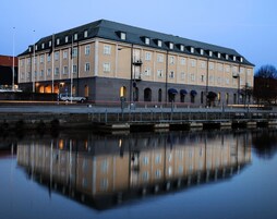 Un edificio grande de varios pisos con fachada simétrica, techo plano y reflejo en el agua de enfrente.