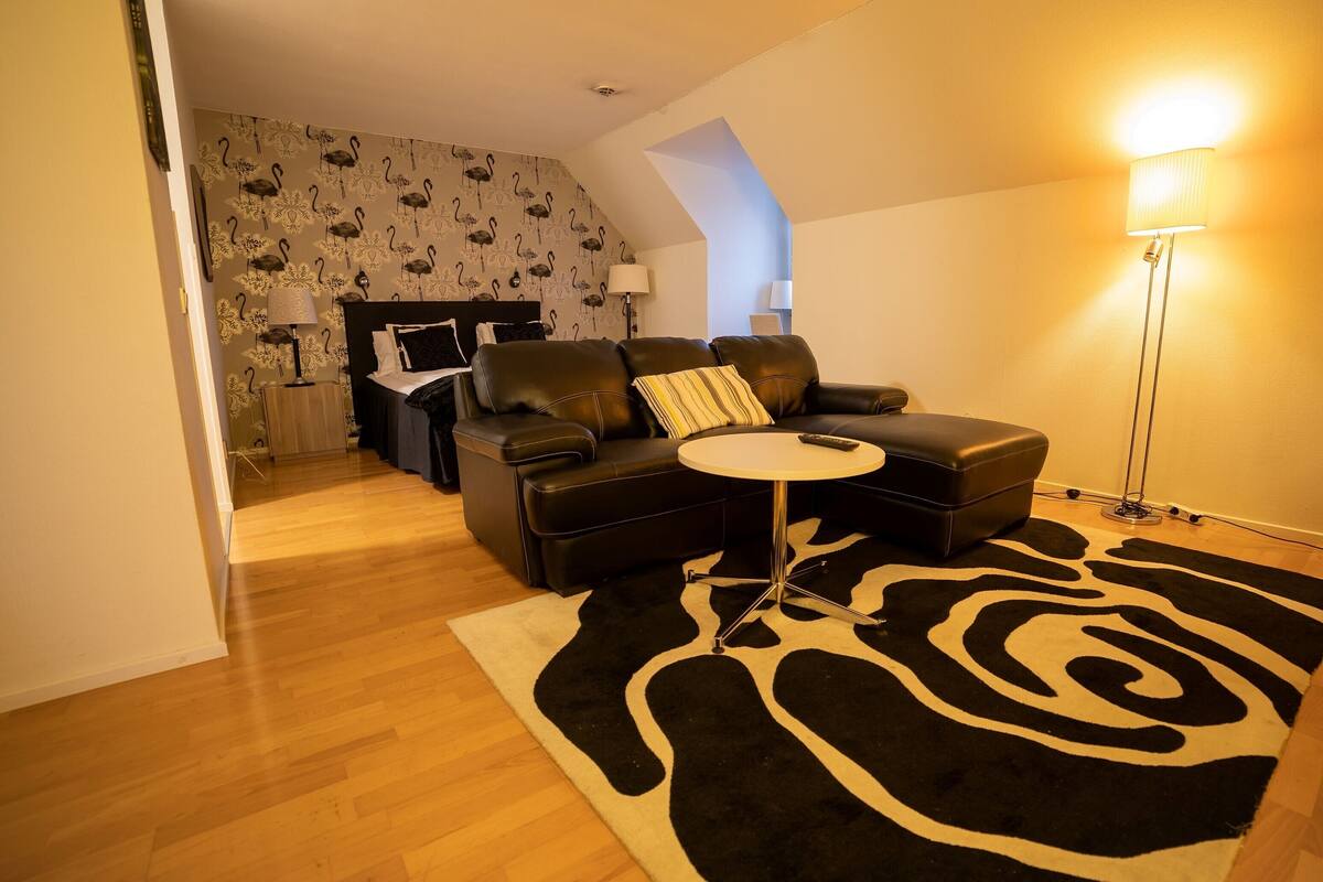 A modern living room with a black leather sofa, a small round table, and a zebra-patterned rug. A bed with a floral wallpaper and a nightstand with a lamp are visible in the background.