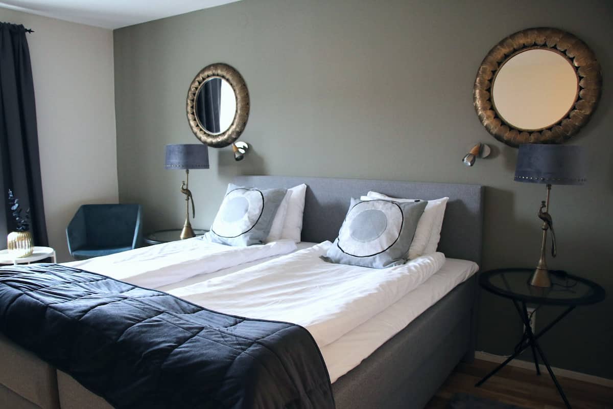 A neatly made bed with a black and white duvet, two bedside lamps, a round mirror, and a small round table with a lamp.