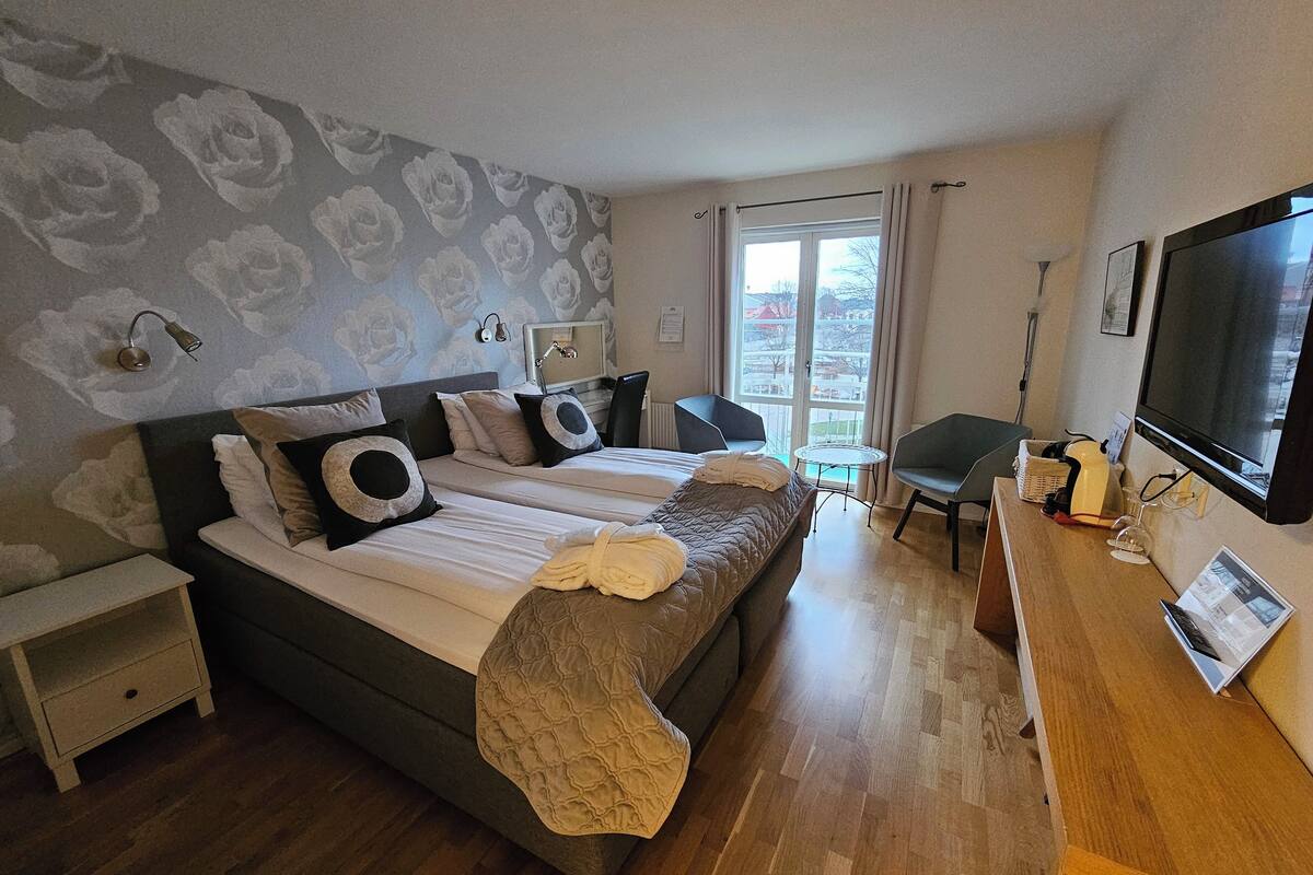 A hotel room with a bed, a desk with a TV, a chair, and a window with a view of buildings.