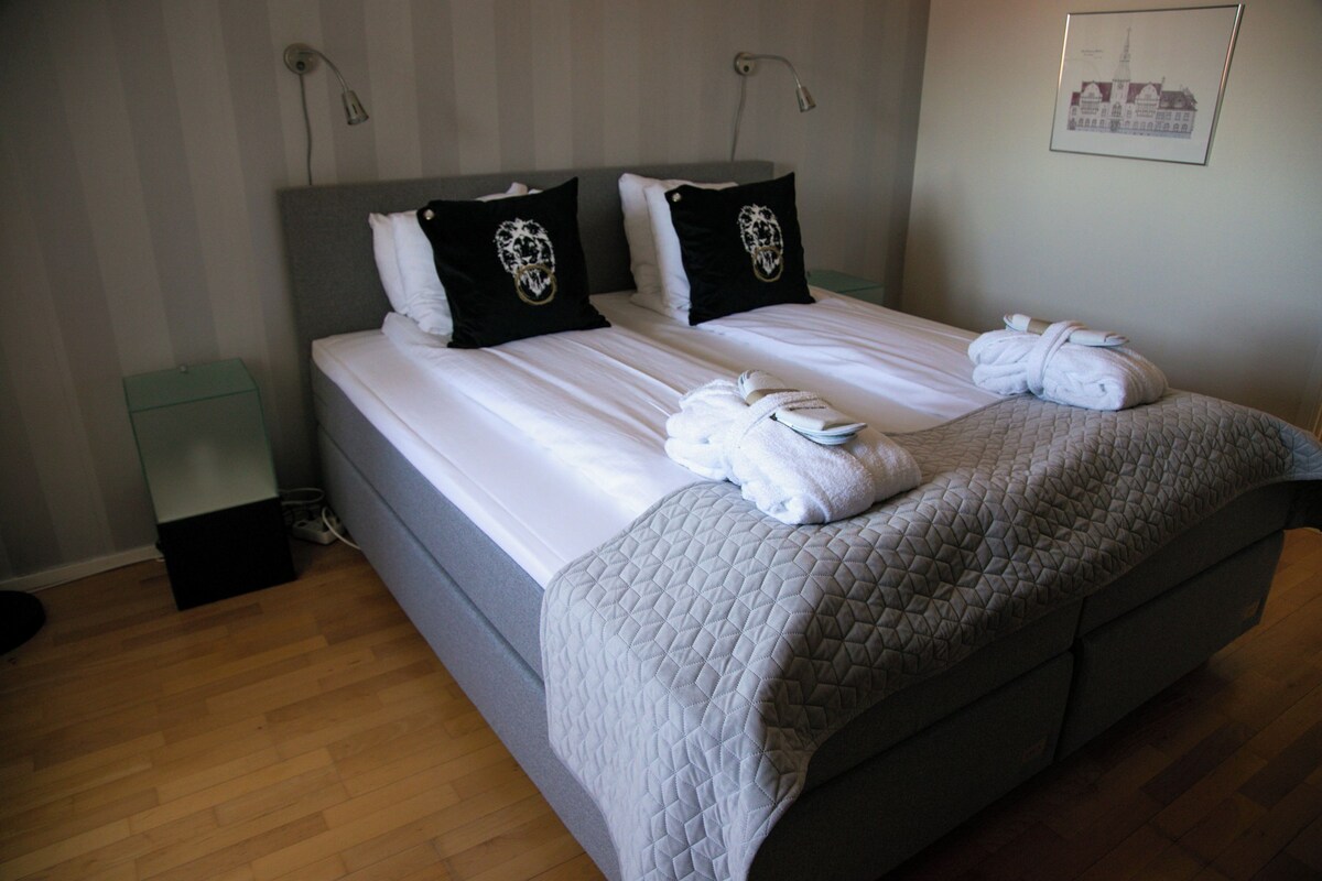 Una cama doble con un edredón gris acolchado, dos almohadas negras con diseño de calavera y dos toallas dobladas sobre la cama.