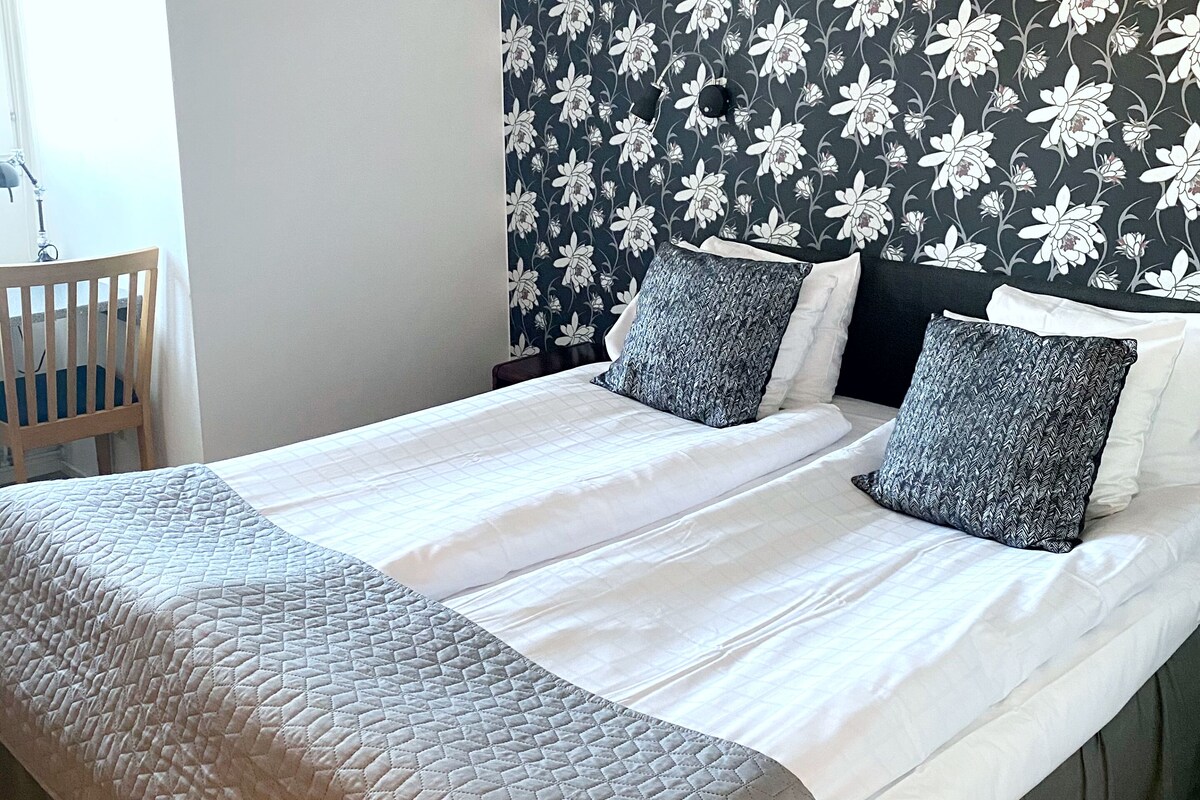 Una cama con colcha blanca acolchada y dos almohadas, una mesita de noche con lámpara y un cabecero estampado con motivos florales.
