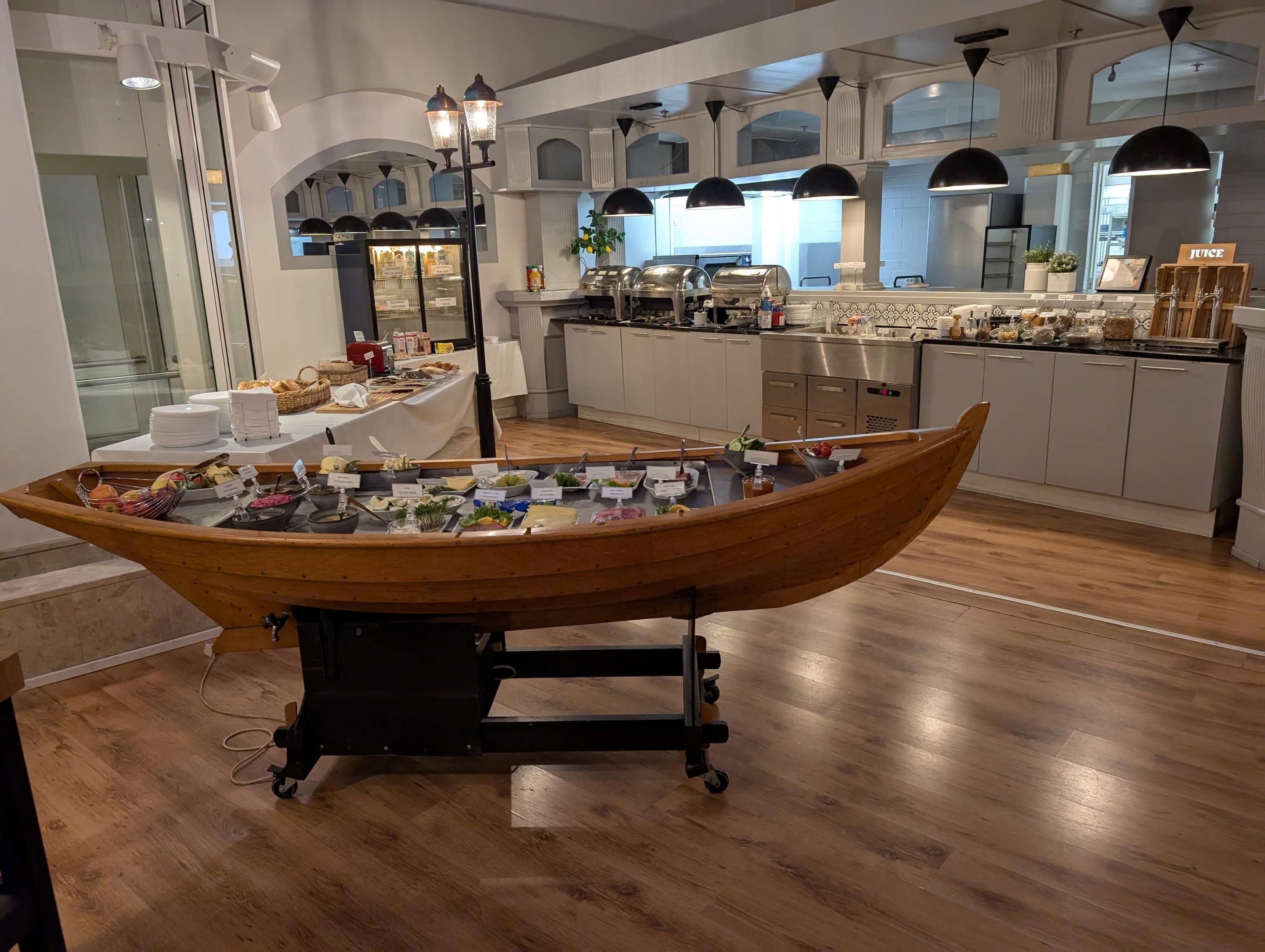 Montaje de bufé con un barco de madera como pieza central, rodeado de diferentes alimentos y un área de cocina al fondo.