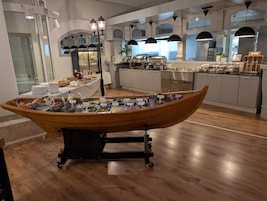 Montaje de bufé con un barco de madera como pieza central, rodeado de diferentes alimentos y un área de cocina al fondo.