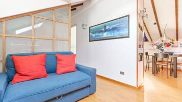 Ein modernes Wohnzimmer mit einem blauen Sofa, roten Kissen, einem Essbereich mit Tisch und Stühlen und einem großen Fenster mit Meerblick.