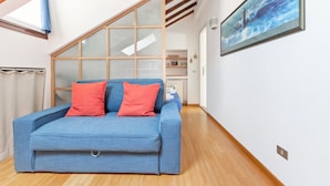 Una habitación con un sofá azul, un separador de vidrio, una obra de arte enmarcada y piso de madera.