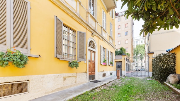 Un estrecho callejón con un edificio amarillo, una puerta de madera y un pequeño jardín con plantas y un árbol.