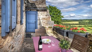 Ein Steinhaus mit einer blauen Tür, einem Tisch mit einer Flasche und Gläsern sowie einem Blumenkasten mit roten Blumen.