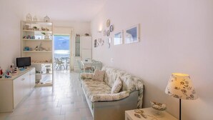 A bright living room with a sofa, a TV, and a lamp. There is a dining area with a table and chairs, and a kitchen with shelves and a TV. - La Casa del Mare close to the beach (Nerano)