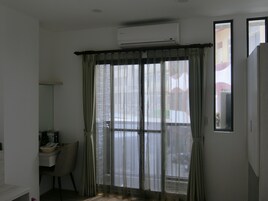 A room with a large window covered by sheer curtains, an air conditioning unit mounted on the wall, a chair, and a small table with items on it.