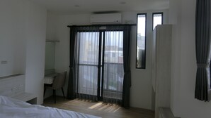 Une chambre d’hôtel avec un lit, un bureau et une chaise. Il y a une fenêtre avec des rideaux, un appareil de climatisation et une vue sur les bâtiments à l’extérieur.