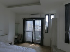 A hotel room with a bed, a desk, and a chair. There is a window with curtains, an air conditioning unit, and a view of buildings outside.