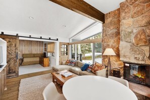 A spacious living area with a stone fireplace, wooden beams, a sofa with colorful cushions, and a dining table.