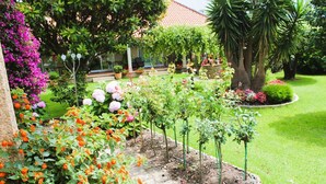 Un jardín bien cuidado con diversas plantas y flores, césped y un edificio con tejado de tejas al fondo.