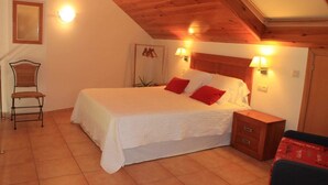 Un dormitorio con techo de madera, una cama con ropa blanca y almohadas rojas, dos lámparas de noche, una silla y una mesita de noche de madera.
