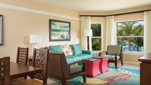 A hotel room with a dining area, a sofa, a chair, a small table, and a large window with a view of palm trees and a pool.