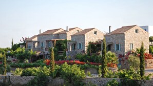 A row of stone houses with a stone wall in front, surrounded by lush greenery and flowers. - Palaeopoli Villas (Kithira)