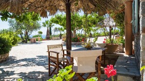 An outdoor seating area with a table and chairs under a thatched roof, surrounded by greenery and a stone pathway. - Palaeopoli Villas (Kithira)