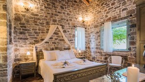 A stone-walled bedroom with a four-poster bed, a nightstand with a lamp, a chair, and a window with curtains. - Palaeopoli Villas (Kithira)
