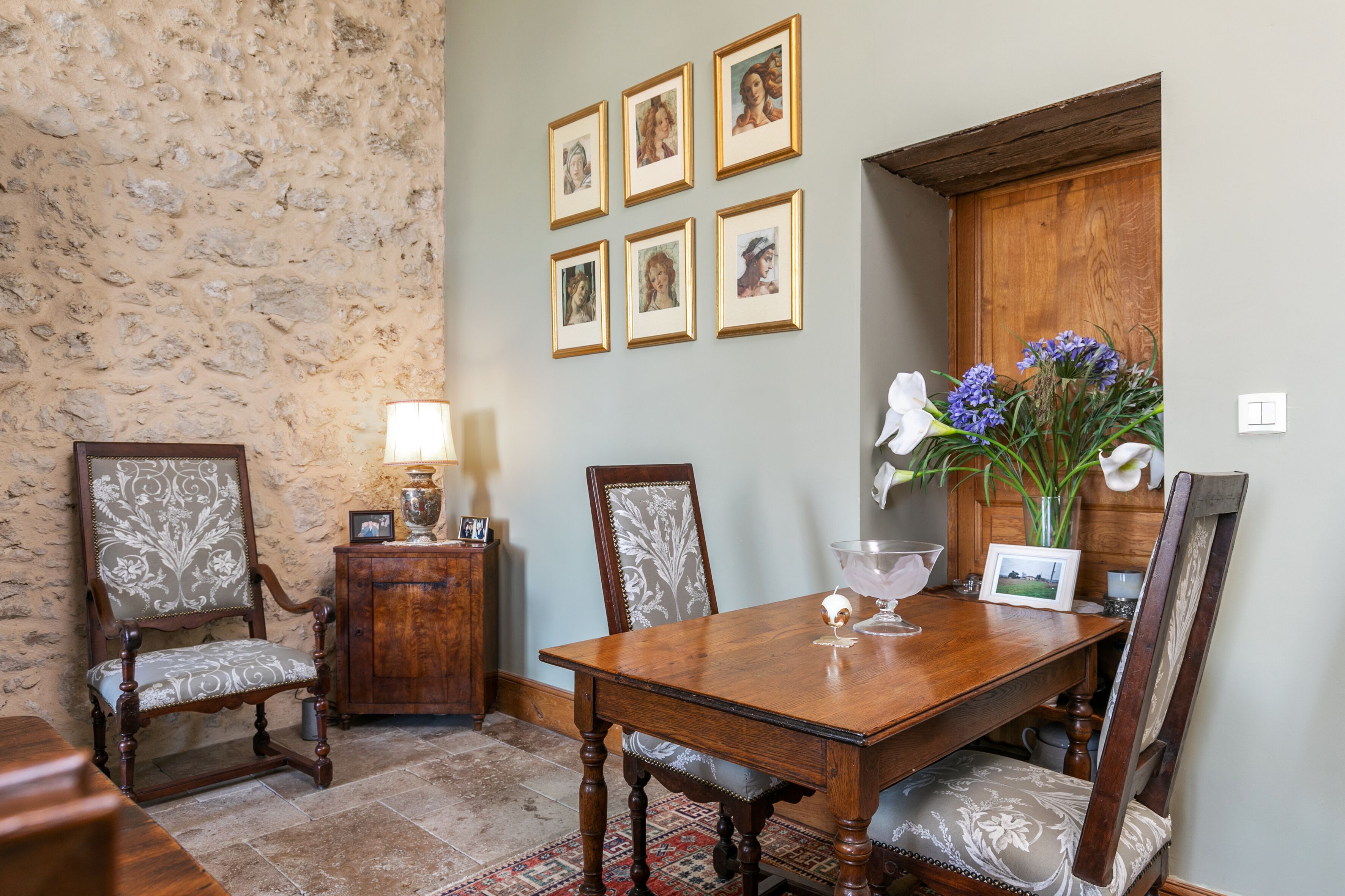 Un comedor con una mesa de madera, sillas y una pared de piedra. Hay cuadros en la pared y un jarrón con flores sobre la mesa.