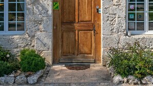 A stone building with a wooden door, a metal decorative panel above the door, and a window on each side. - Domaine de Rambeau (Castelmoron-sur-Lot)