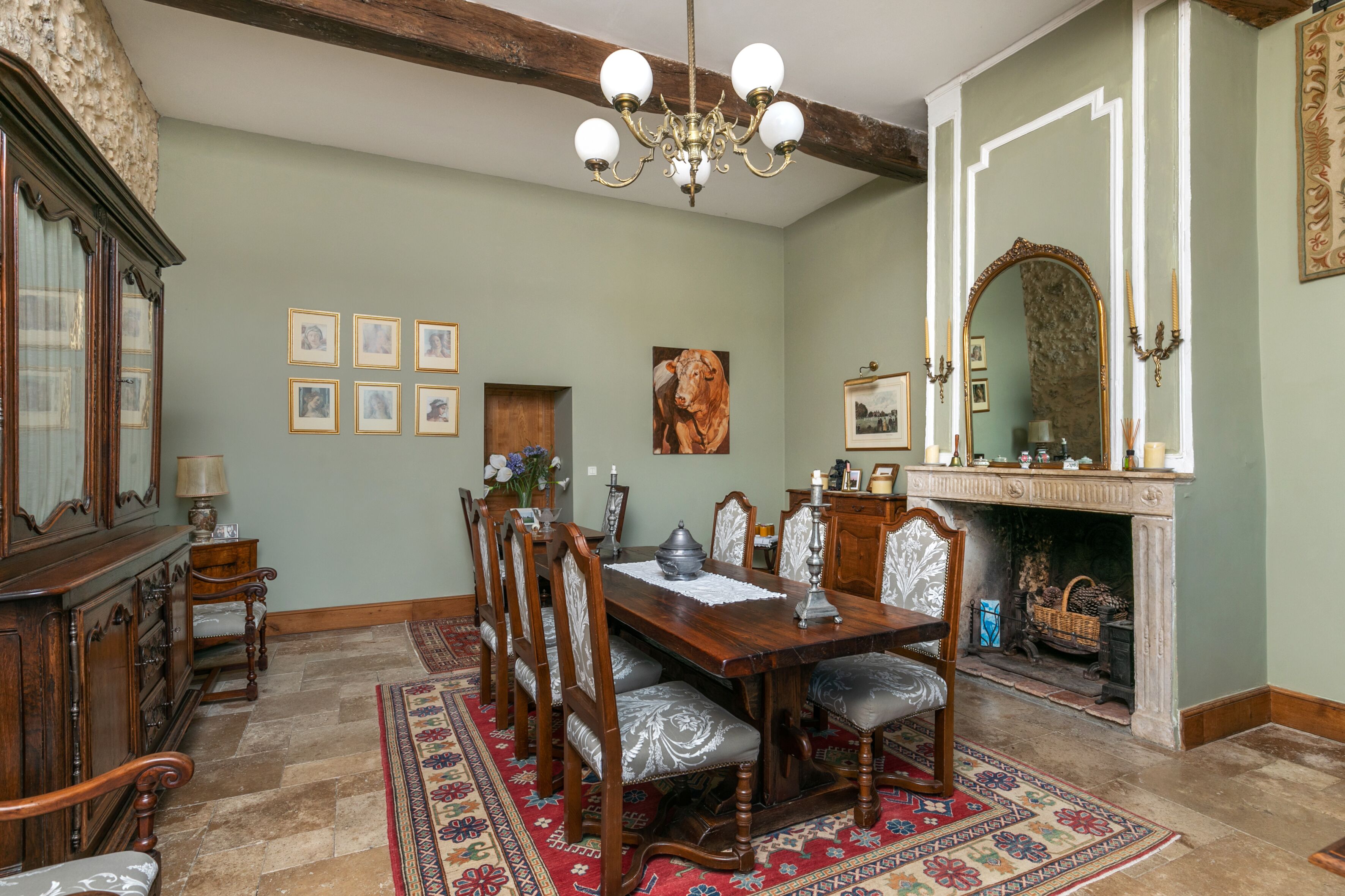 Un comedor con una mesa de madera, sillas, una araña, chimenea y cuadros en la pared.