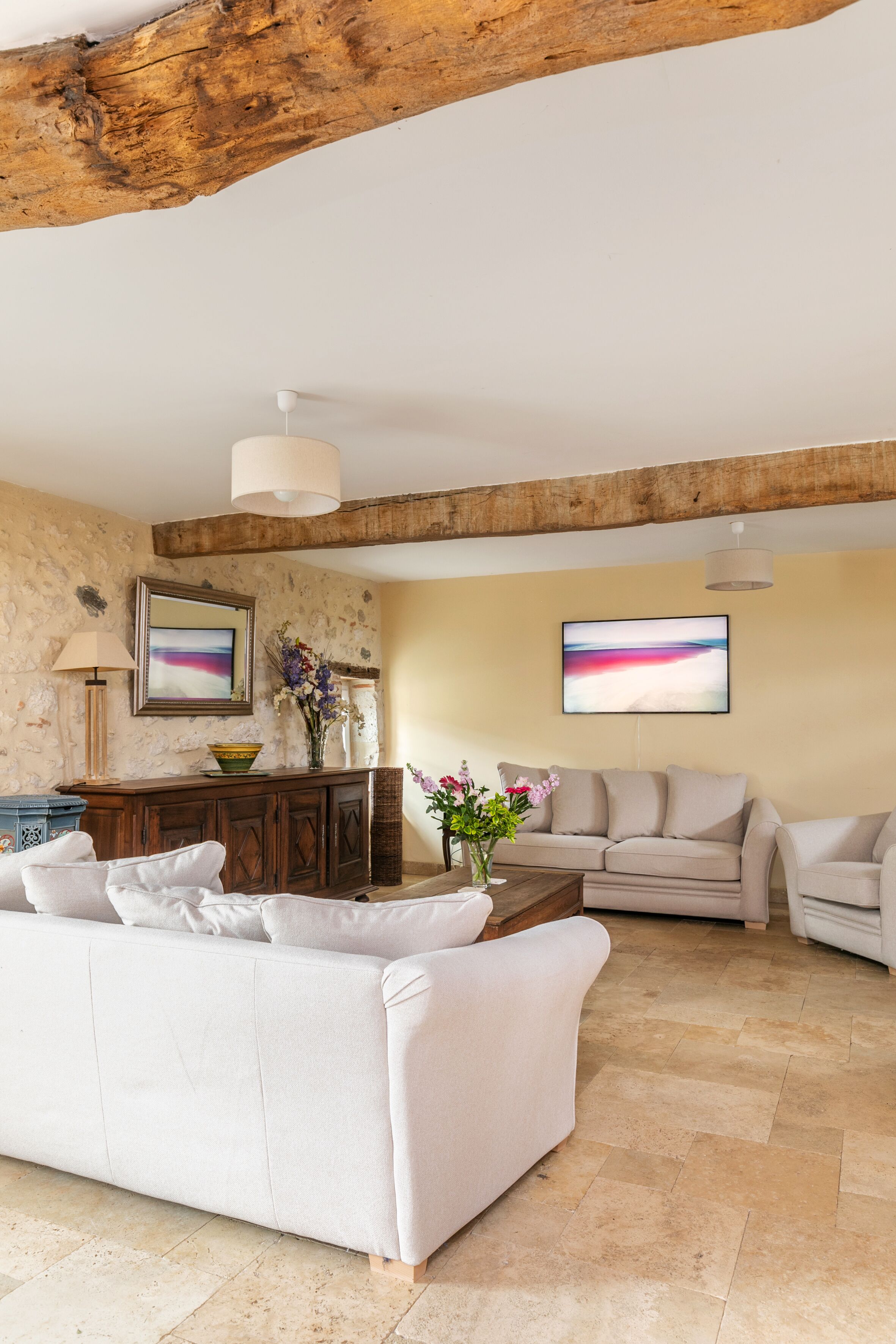 A spacious living room with a white sofa, a wooden coffee table, and a beige couch. There are two framed artworks on the wall and a vase with flowers on the coffee table.